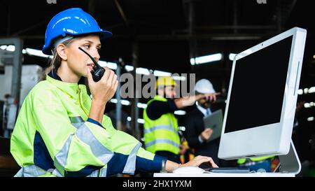 Ingegnere meccanico donna che tiene radio industria di lavoro indossando gilet di sicurezza riflettente e casco blu, ispettore controllo in piedi macchina industriale Foto Stock