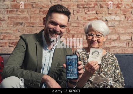 figlio caucasico di mezza età seduto su un divano con la madre anziana pensionato che guarda la macchina fotografica e mostra la galleria fotografica e le foto stampate. Foto di alta qualità Foto Stock