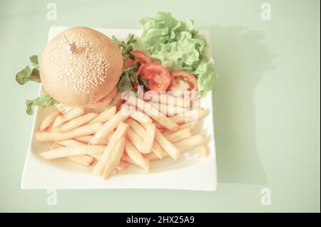 Hamburger con sesamo e fritte francesi con verdure servite su un piatto bianco su un tavolo verde chiaro sullo sfondo. Vista dall'alto. Disposizione piatta. Primo piano. Foto Stock