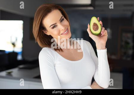 Felice giovane 30s donna caucasica che mostra metà avocado in cucina Foto Stock