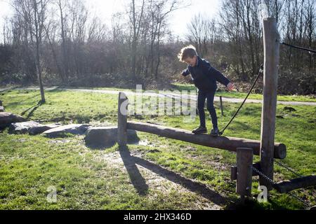 Un giovane ragazzo perde quasi il suo equilibrio su un fascio di legno su un percorso di ostacoli in un parco, in sole di primavera brillante, in una foresta a Diemen, nei Paesi Bassi. Foto Stock