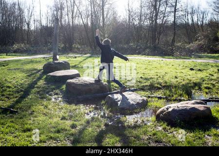 Un giovane ragazzo salta sulle pietre di un percorso a ostacoli in un parco, al sole della sorgente luminosa, in una foresta a Diemen, nei Paesi Bassi. Foto Stock