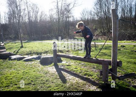 Un giovane ragazzo si bilancia su una trave di legno su un percorso ad ostacoli in un parco, al sole di primavera, in una foresta a Diemen, nei Paesi Bassi. Foto Stock