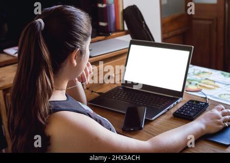 vista posteriore della giovane donna caucasica a casa guardando il suo schermo laptop con una mano sul mouse e un telefono cellulare sulla tastiera. Foto Stock