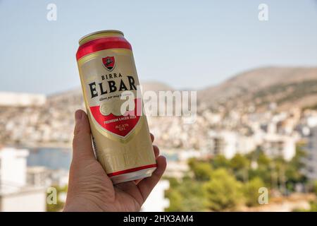 Saranda, Albania - 29 luglio 2021: La mano dell'uomo bianco tiene una lattina di birra albanese Elbar in primo piano con paesaggio urbano sullo sfondo. Foto Stock