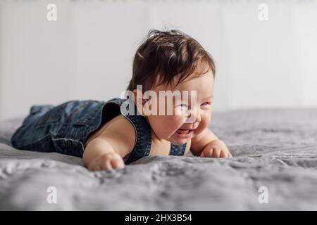 Ritratto del bambino grazioso che indossa il romper del denim che giace sulla coperta grigia nel paese. Piangendo bambino strisciando sul letto in camera da letto, sfondo bianco Foto Stock