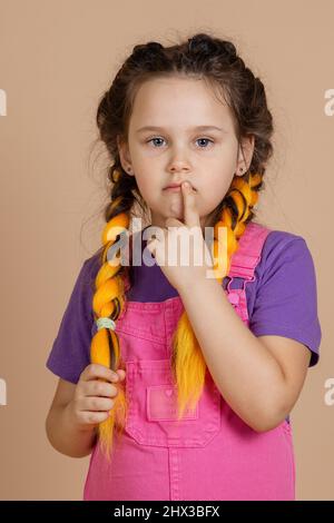 Pensive piccolo, femmina bambino con occhi seri guardando la fotocamera con canekalon giallo trecce toccare la bocca con un dito in rosa tuta e. Foto Stock