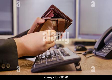 Un uomo d'affari con un portafoglio vuoto senza soldi nelle sue mani sta lavorando su una tastiera del calcolatore ad una scrivania dell'ufficio, primo piano Foto Stock