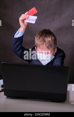 Un ragazzo in un abito da scuola con una maschera medica sul viso e carte di credito al computer, spazio copia su uno sfondo scuro studio Foto Stock