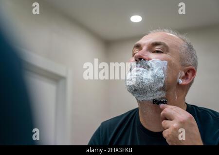 Uomo maturo dai capelli grigi che restyling la sua barba in casa utilizzando il rasoio Foto Stock