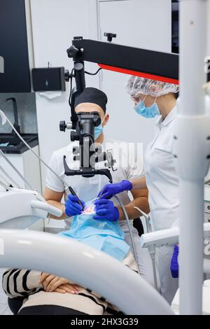 Un dentista di successo guarda i denti del paziente con un microscopio dentistico e tiene gli strumenti dentali vicino alla bocca. L'assistente aiuta il medico Foto Stock