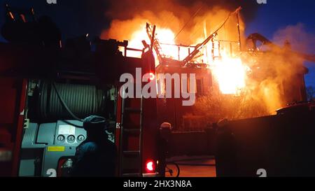 Il tetto della casa è sul fuoco. L'edificio residenziale bruciare, villaggio. I vigili del fuoco hanno messo fuori un fuoco. Vigili del fuoco che mette fuori la fiamma. Camion antincendio, fari e pannello, sirena, segnale, allarme. Foto Stock