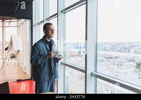 Un giovane di aspetto europeo in un abito informale beve caffè e guarda fuori dalla finestra Foto Stock