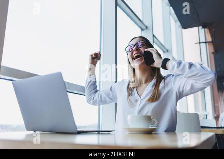 Una giovane donna manager in occhiali lavora a un computer portatile e gesticula emotivamente con le mani mentre parla al telefono, è seduta alla sua scrivania Foto Stock