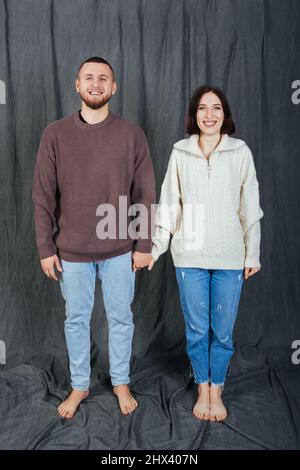 Giovane e bella coppia shyly che tiene le mani su una prima data. Tessuto grigio background. Indossano jeans casual e maglioni. Foto Stock