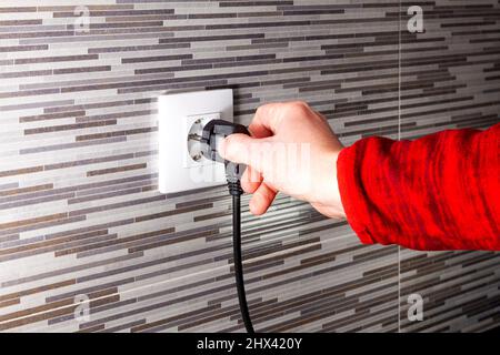 La mano di un uomo sconosciuto che collega una spina europea nera a una presa elettrica installata in una parete piastrellata. Il manicotto dell'abbigliamento è rosso. Foto Stock