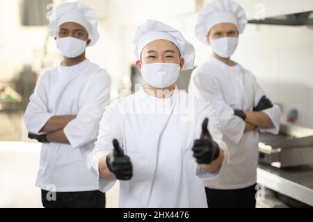 Ritratto di tre cuochi chef ben vestiti con diverse etnie in piedi insieme in cucina ristorante. Chef asiatico mostra OK segno, latino e europeo ragazzi sullo sfondo. Cuochi che indossano maschere per il viso pronti a cucinare durante la pandemia Foto Stock