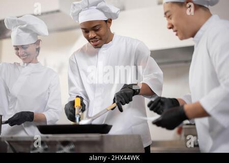 Multirazziale di cuochi professionali in uniforme preparare i pasti per un ristorante in cucina. Chef asiatico che gestisce il processo, ragazzo latino e cucina europea friggere. Lavoro di squadra e duro lavoro al ristorante, occupazione professionale Foto Stock