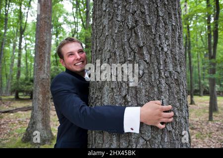 SORRIDENTE GIOVANE UOMO D'AFFARI ABBRACCIANDO VECCHIO ALBERO DI PINO DI CRESCITA IN FORESTA SETTENTRIONALE MISTA TEMPERATA Foto Stock