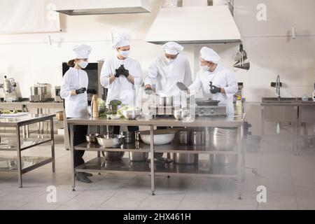 Multirazziale di cuochi in bianco uniforme cucina insieme in cucina ristorante. Concetto di lavoro di squadra e duro lavoro al ristorante. Cuochi che indossano le maschere di faccia per impedire la diffusione del virus Foto Stock