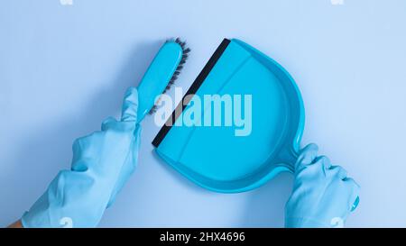 Mani in guanti di gomma blu che tengono scopa e fertilizzante su sfondo blu pastello. Strumenti di pulizia e concetto di lavori domestici. Disposizione piatta Foto Stock