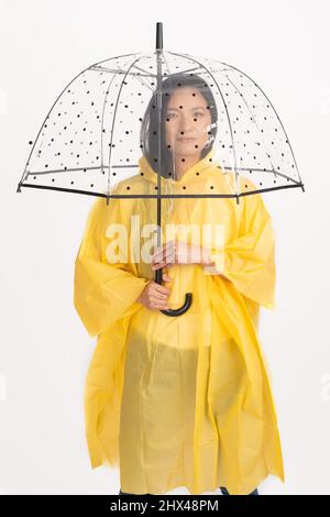 Foto studio di bella donna asiatica con lunghi capelli neri indossando impermeabile giallo e nascondendosi sotto trasparente con ombrello nero puntini con copia spa Foto Stock