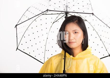 Foto studio di bella donna asiatica con lunghi capelli neri indossando impermeabile giallo e nascondendosi sotto trasparente con ombrello nero puntini con copia spa Foto Stock