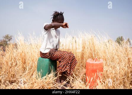 Bambina esausta seduta su un barattolo di plastica in un campo asciutto nella macchia africana che soffre di calore bruciante e il suo duro lavoro; Child la Foto Stock