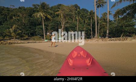 Giovane donna e occhiali da sole tira canoe rosa in plastica con pagaia per azzurro acqua di mare dalla spiaggia con palme. Inizio della gita in barca, imbarco. Viaggiare in paesi tropicali Foto Stock
