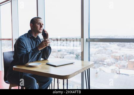 Happy man con chiamate di caffè tramite smartphone mentre il computer portatile in remoto lavora alla scrivania, un allegro freelance che si gode la pausa caffè mentre lavora remotel Foto Stock