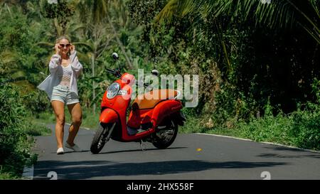 Donna su scooter rosso in abiti bianchi guida su strada forestale. Gita danzante. Una ragazza caucasica turista in occhiali da sole danza, relax, riposo vicino moto. Asia Thailandia giro turismo. Noleggio moto. Foto Stock