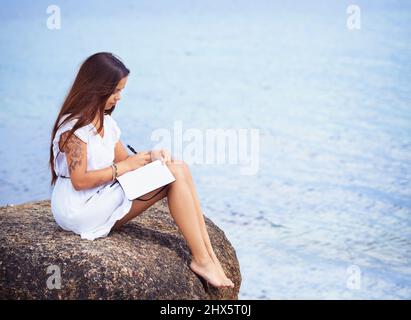 Approfondimenti profondi circondati dalla natura tranquilla. Scatto a tutta lunghezza di una giovane donna tatuata gorgeous seduta su una scrittura di roccia in un diario. Foto Stock