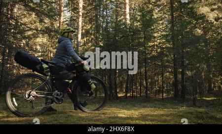 La donna viaggia in bicicletta su terreni misti con bikepacking in bicicletta all'aperto. Il viaggio del viaggiatore con le borse della bicicletta. Bikepacking elegante, bici, abbigliamento sportivo in verde nero. Parco magico della foresta. Foto Stock