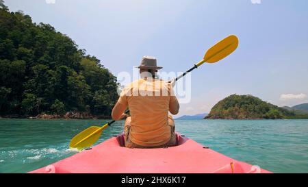 Giovane uomo con occhiali da sole e cappello ranghi rosa canoe di plastica lungo il mare contro verdi isole collinari con giungle selvatiche. Viaggiare in paesi tropicali. Il ragazzo forte sta navigando in kayak nell'oceano, vista posteriore. Foto Stock