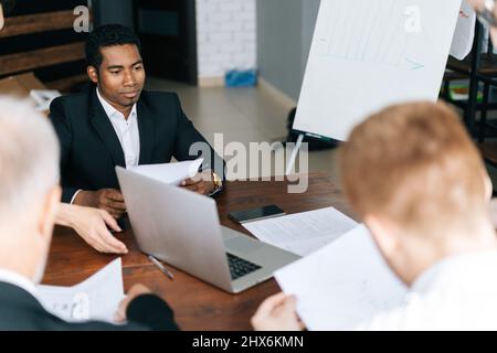 Vista ad alto angolo di uomini d'affari multietnici focalizzati che controllano il documento d'accordo prima di firmarlo seduti alla scrivania in un ufficio moderno. Foto Stock