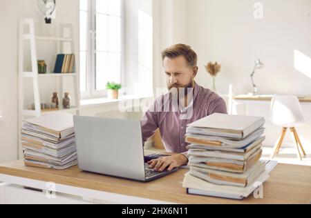 Serio contabile seduto alla sua scrivania d'ufficio con documenti e di lavoro sul suo laptop Foto Stock