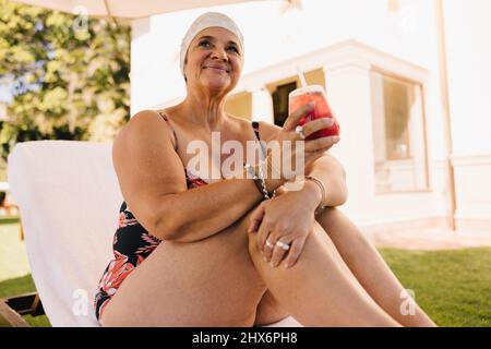 Donna anziana spensierata che si rilassa su un lettino con un cocktail in mano. Donna anziana che si gode la sua vacanza estiva in un resort termale di lusso. Felice matu Foto Stock