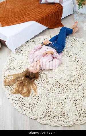 Una donna si trova su un tappeto vicino al letto, un tappeto fatto in casa a maglia fatto di fili spessi, fatto a mano: Un tappeto fatto a mano fatto di fili beige giace sul fl Foto Stock