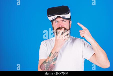 Uomo con barba e baffi con occhiali VR, sfondo blu. Ragazzo con occhiali VR o display montato sulla testa. Hipster su faccia pensierosa che punta a. Foto Stock