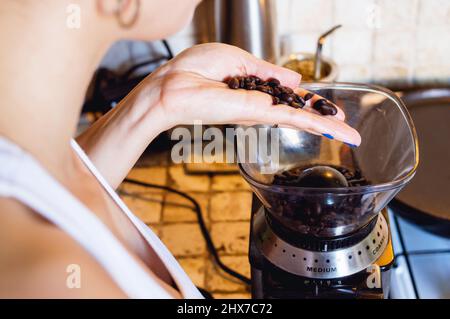 primo piano della giovane donna caucasica in cucina versando caffè in chicchi in un macinacaffè elettrico sul bancone, stile di vita e concetto di industria del caffè Foto Stock