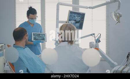 Il dentista punta alla radiografia dentaria su tablet digitale, spiegando la diagnosi al paziente con carie. Medico che mostra i risultati della scansione a raggi X per l'uomo in dolore per fare la procedura chirurgica nel cabinet. Foto Stock