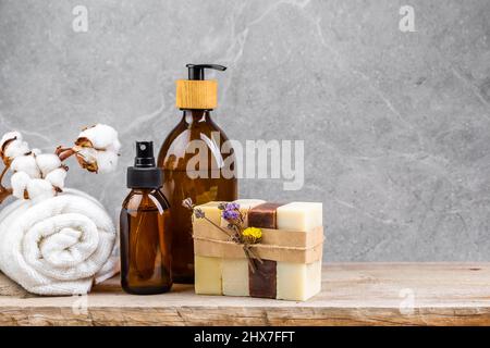 Tavolo da bagno con asciugamano bianco e ramo di fiori di cotone, bottiglie di vetro scuro con prodotti per la cura della pelle liquida e saponette naturali fatte a mano in st Foto Stock