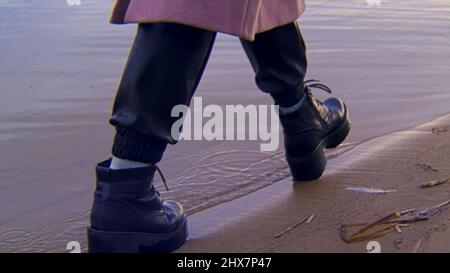 Primo piano di stivali neri in pelle di sesso femminile che camminano su una spiaggia sabbiosa e il bordo d'acqua. Riprese in stock. Dettagli di una giovane donna che cammina vicino al fiume. Foto Stock