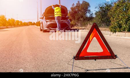 Auto con problemi e un triangolo rosso per avvisare gli altri utenti della strada. Conducente maschio in piedi vicino a un'auto rotta con cofano aperto. Foto Stock