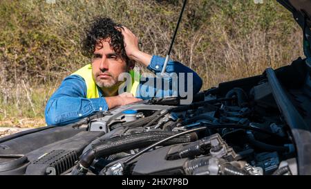 Faccia di conducente maschio infelice vicino ad una macchina rotta con cofano schioccato ispezionare il suo motore del veicolo. Stanco e stressato uomo che tiene testa a portata di mano un Foto Stock