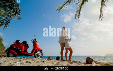 Viaggio su strada in scooter. Bella coppia su moto rossa in abiti bianchi sulla spiaggia di sabbia. Matrimonio appena sposato persone che camminano vicino a palme tropicali, mare. Ho Foto Stock