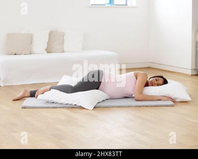 Donna incinta sdraiata sul pavimento sul tappeto in soggiorno con testa e gambe sostenute da grandi e confortevoli cuscini durante la prima fase del travaglio Foto Stock