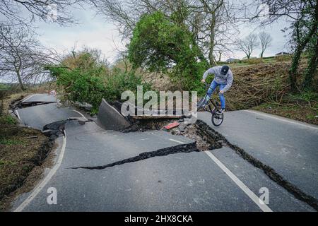 Un uomo su una BMX salta sopra un gap nel asfalto rotto su una sezione del B4069 vicino Lyneham nel Wiltshire, che è ancora in movimento e in cambiamento di forma, a seguito di una striscia di terra nel mese di febbraio. Data foto: Giovedì 10 marzo 2022. Foto Stock