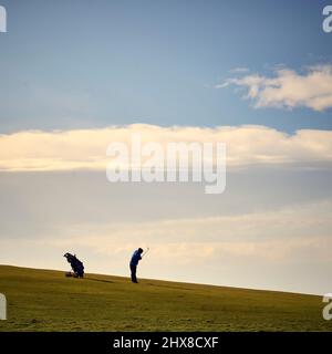 Solitario golfista che gioca colpo sul campo da golf in pendenza contro la luce Foto Stock
