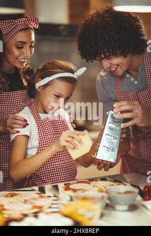 I genitori felici e la loro figlia stanno preparando il pasto insieme nella cucina. La bambina e suo padre grattugiano il formaggio e lo mettono sulla pizza Foto Stock